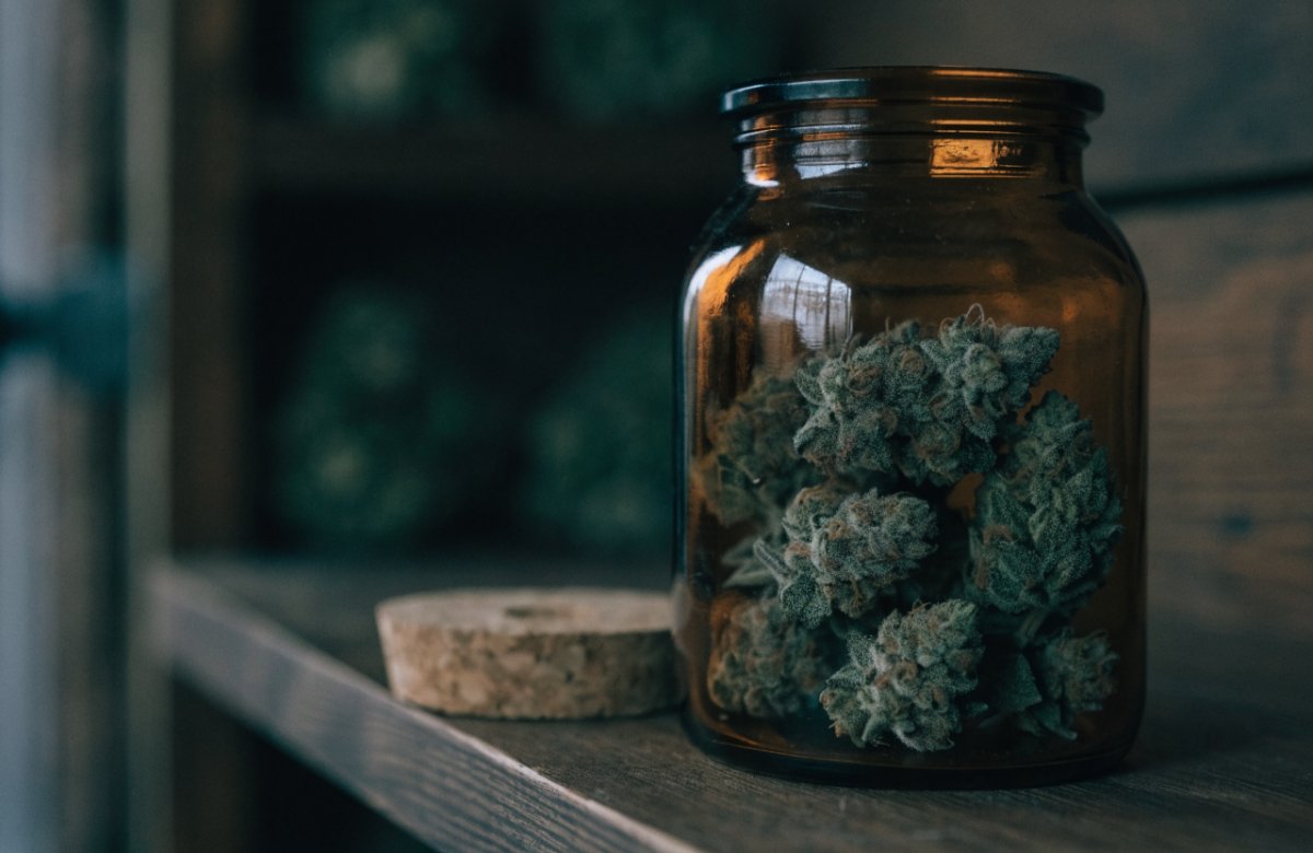 Cured cannabis buds stored in an amber glass jar on a dark wooden shelf, the airtight, cool and dark storage that keeps weed fresh