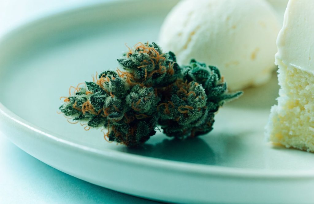 Dense forest-green Ice Cream Cake cannabis buds with orange hairs on a pale plate beside vanilla ice cream and a wedge of cake