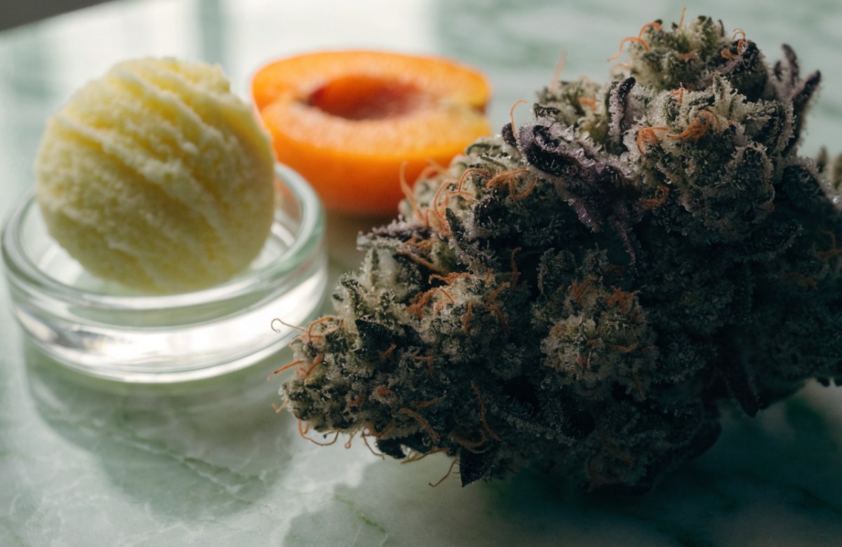 A frosty olive-green and purple Jealousy cannabis bud with orange pistils on pale green marble beside a scoop of sherbet in a glass dish and a halved apricot